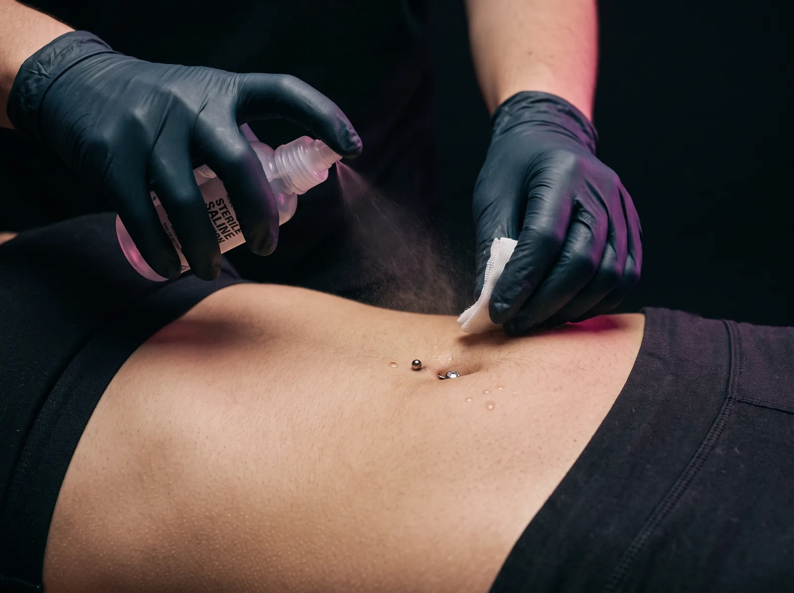 Gloved hands performing sterile saline aftercare on a fresh navel piercing