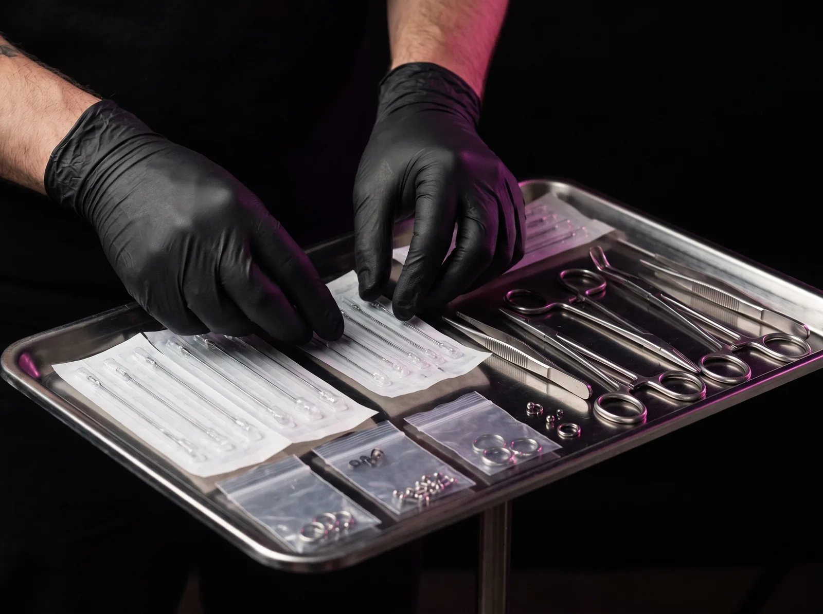 Gloved hands arranging a sterile single-use needle and jewelry tray before a piercing session